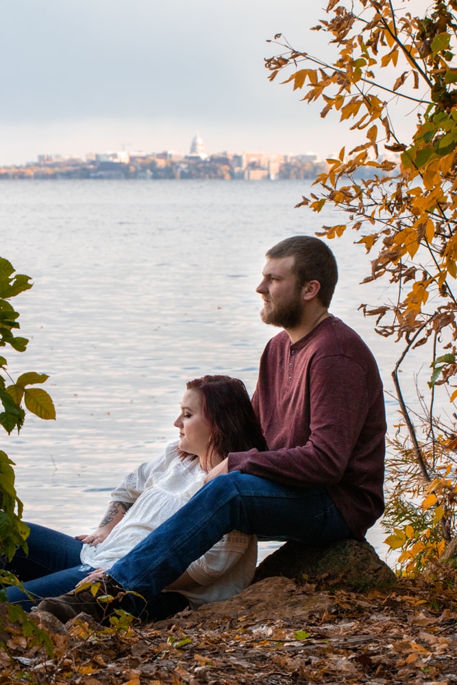 Couple sharing intimate autumn moment by lake with Madison, Wisconsin skyline during golden hour portrait session