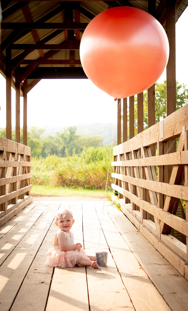 Joyful baby in tutu sits on wooden bridge with coral balloon, milestone portrait session Madison Wisconsin
