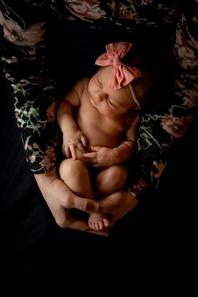 Peaceful newborn baby with coral bow resting in parent's hands, Madison Wisconsin newborn portrait photography