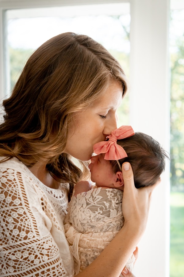 Tender mother kisses sleeping newborn baby with coral bow in bright Madison Wisconsin portrait session